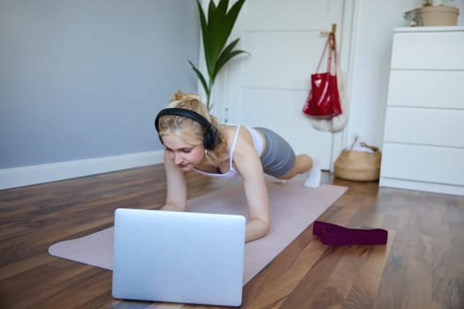 Portrait of young athletic woman in wireless headphones, standing in plank, following online workout video, using laptop to join training session on the internet.