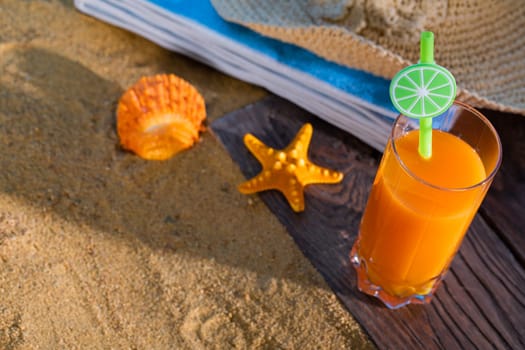 Fresh orange fruit juice stands in a glass with a straw. A beach towel lies on the sandy beach.