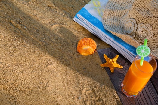 Fresh orange fruit juice stands in a glass with a straw. A beach towel lies on the sandy beach.