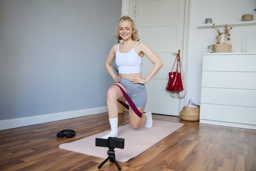 Portrait of young athletic woman recording home workout video, shooting content for sport fitness vlog, using resistance band and digital camera.