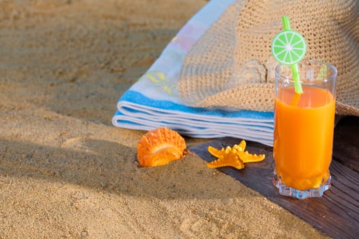 Fresh orange fruit juice stands in a glass with a straw. A beach towel lies on the sandy beach.