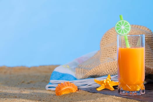 Fresh orange fruit juice stands in a glass with a straw. A beach towel lies on the sandy beach. Blue sky.