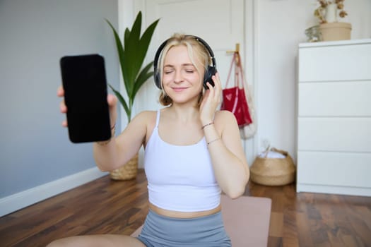 Portrait of smiling, beautiful young woman, showing her smartphone screen, wearing headphones, looking satisfied with sound quality.