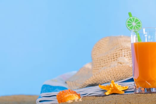 Fresh orange fruit juice stands in a glass with a straw. A beach towel lies on the sandy beach. Blue sky.