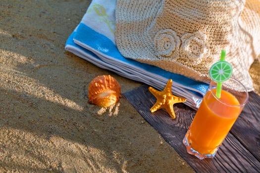 Fresh orange fruit juice stands in a glass with a straw. A beach towel lies on the sandy beach.