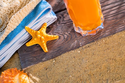 Fresh orange fruit juice stands in a glass with a straw. A beach towel lies on the sandy beach.