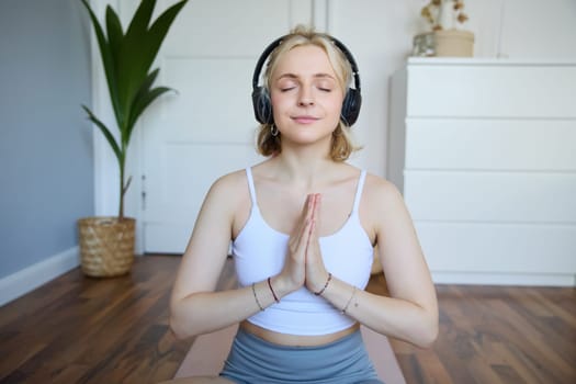 Portrait of young relaxed woman, sits in room in headphones, clasp hands together, meditated, listens to yoga music.