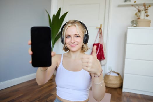 Portrait of young fitness instructor in headphones, showing smartphone screen and thumbs up, recommending app for home workout.