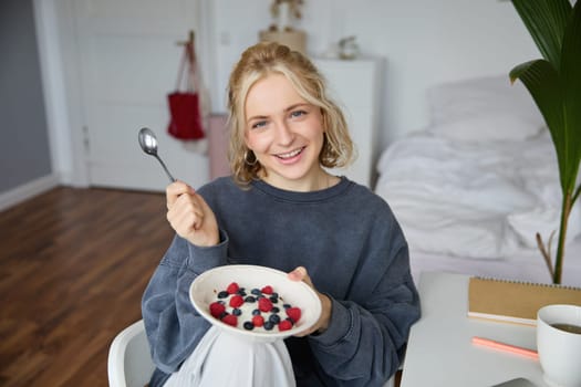 Portrait of smiling blond woman, eating breakfast, holding bowl and spoon, sitting in bedroom, looking happy at camera. Lifestyle concept