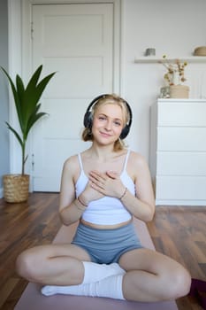 Vertical portrait of beautiful young woman in headphones, sits on yoga mat, meditating, holding hands on her chest and smiling at camera.
