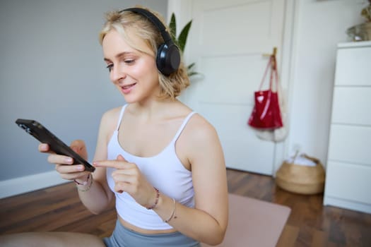 Close up portrait of athletic young woman, holding mobile phone and wearing wireless headphones, workout at home, using sport app during fitness training.