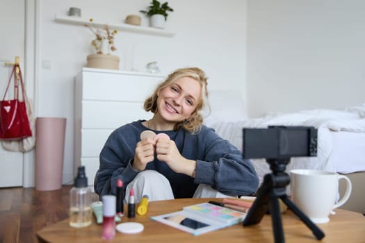 Portrait of smiling young woman, sitting in front of digital camera in cosy set up, recording video for lifestyle vlog, chatting about makeup and beauty products.