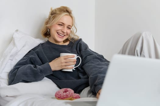Portrait of candid, happy young woman lying in bed, looking at laptop screen, holding cup of tea and eating doughnut, staying at home on weekend, spending quite time alone.