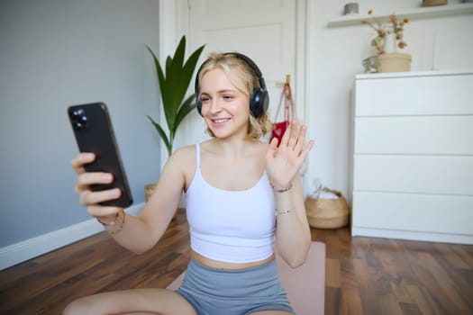 Portrait of young woman with smartphone and headphones, waving hand at mobile phone camera, live streaming during workout. Wellbeing and sport concept