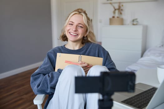 Portrait of smiling, charismatic young woman, content creator, records video on digital camera, laughing and looking happy, using journal, writing in notebook.
