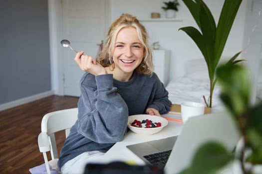 Image of laughing woman sitting in front of laptop in her room, eating breakfast, holding spoon and bowl in hand, watching videos online while having a snack.