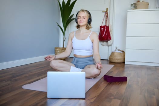 Portrait of concentrated woman in headphones, meditating, listening to calm music and practice yoga, follow instructions on laptop, sitting on rubber mat.