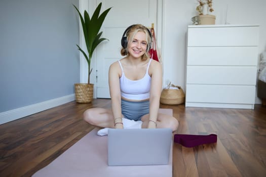 Portrait of woman during workout, sitting on yoga mat with resistance band, listening to video instructions on laptop, wearing wireless headphones, repeating exercises.