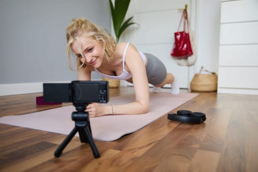 Young woman, personal fitness instructor records video of herself standing in a plank, using rubber yoga mat and digital camera at home.