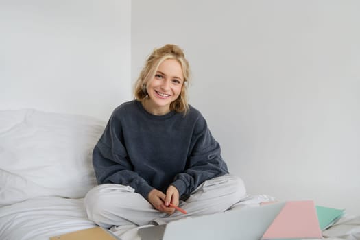 Portrait of young beautiful woman sitting on bed with laptop and notebooks, working from home, freelancing. Female student studying online, video chats, connects to lesson remotely.