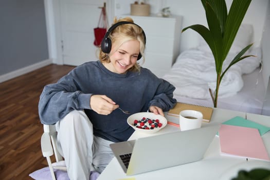 Portrait of smiling young blond woman in headphones, sitting in room, watching movie on laptop, eating breakfast and drinking tea, having lunch in front of computer screen.