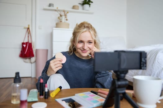 Image of young woman, vlogger records video on digital camera, shows beauty products, recommending makeup for audience on social media, sits on front in a room.