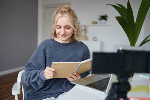 Portrait of young woman with notes, working from home, recording video on digital camera, live streaming from her room, reading from notebook and smiling.