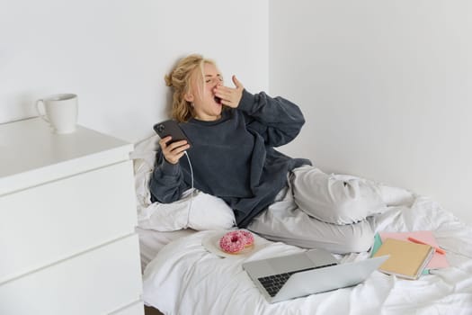 Portrait of young woman spending lazy weekend at home, lying in bed with smartphone and laptop, eats doughnut and yawns, feels sleepy or bored.