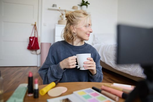 Image of young woman, makeup vlogger, sitting in bedroom with digital camera, drinking tea and talking, creating lifestyle video, social media content.