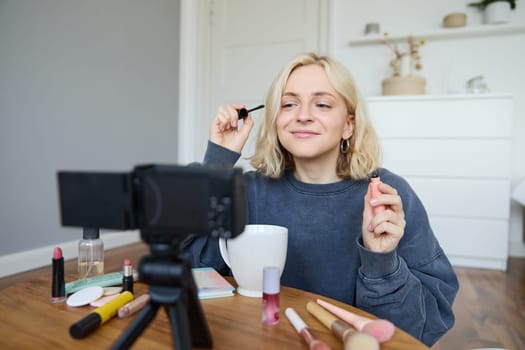 Image of cheerful, beautiful young lifestyle blogger, woman sitting on floor and recording video about makeup, holding mascara, making lifestyle content for her social media account and followers.