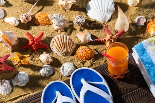 On the sea beach there is a blue towel and beach slippers and next to it in a tall glass is poured fresh fruit juice.