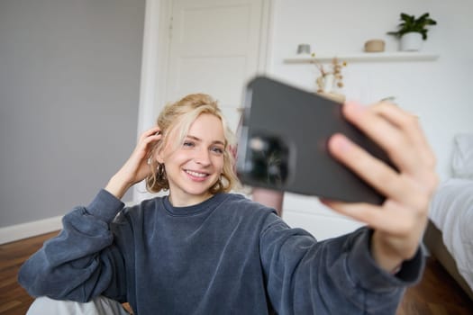 Portrait of young woman, social media influencer, taking selfies in her room, sitting on floor, holding smartphone and posing for a photo.
