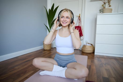Young and sporty woman working out at home, doing fitness training alone on yoga mat, listens to music in wireless headphones.