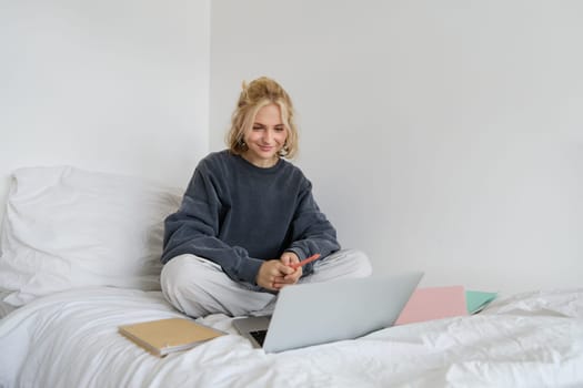 Portrait of happy blond woman, freelancer working from home, sitting on bed with laptop and notebooks. Student doing homework in bedroom, connects to online class via video chat.