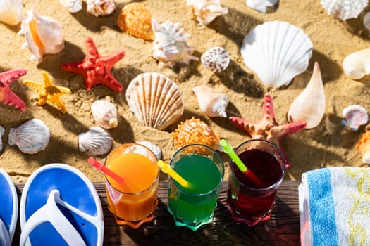 On the sea beach there is a blue towel and beach slippers and next to it in a tall glass is poured fresh fruit juice.