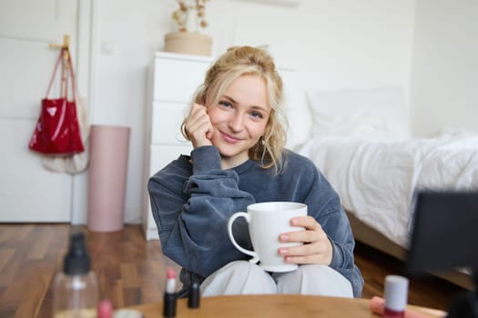 Close up portrait of beautiful blond woman, girl sits in her room, does makeup, records video for social media, drinks tea, holds mug and smiles at camera.