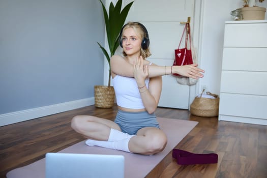 Portrait of beautiful, athletic woman at home, doing workout, watching fitness instructor video on laptop, wearing headphones, stretching her arms.