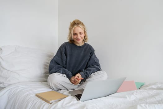 Portrait of young female student, woman studying online, e-learning on her laptop, sitting on bed with notebook and looking at screen, video chats, connects to a self-paced course.