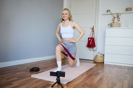 Portrait of fitness instructor, young sporty vlogger woman doing exercises on camera, shooting video about workout, using resistance band and doing sit-ups at home in empty room.