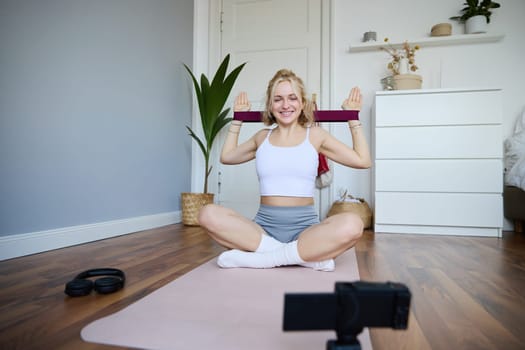 Portrait of young athletic woman recording home workout video, shooting content for sport fitness vlog, using resistance band and digital camera.