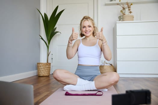 Healthy and fit fitness blogger, recording vlog for wellbeing social media account, sitting on yoga mat and showing thumbs up, using digital camera to create content, following script of laptop.