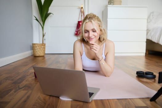 Portrait of young fitness woman, doing workout at home, looking at her gym instructor on laptop screen, doing online training session.