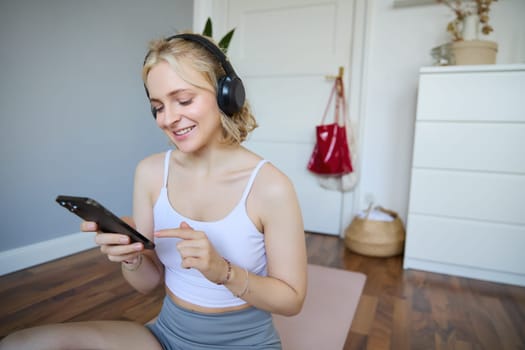 Close up portrait of blond young woman choosing fitness podcast, looking at her mobile phone, checking workout app on smartphone, wearing wireless headphones.