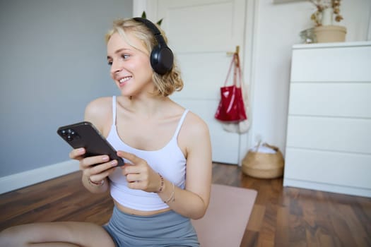 Portrait of smiling, healthy young woman in headphones, holding smartphone, working out at home, using fitness application on mobile phone. Wellbeing and sport concept