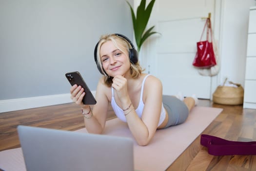 Portrait of young fitness woman, choosing workout on laptop, wearing headphones and using mobile phone exercise app. Sport and lifestyle concept