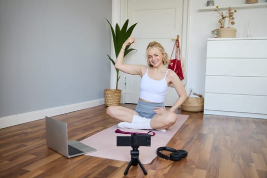 Young female athlete, fitness instructor woman sits on floor rubber mat, recording video on digital camera, showing how to workout, explaining exercises.