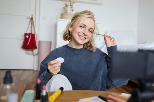 Portrait of smiling young woman applies makeup, shows beauty tutorial on video, records vlog on digital camera in her room, recommends cosmetic products.