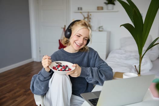 Portrait of smiling beautiful woman, sitting in room with breakfast, eating and watching tv show on laptop, laughing and looking at screen.