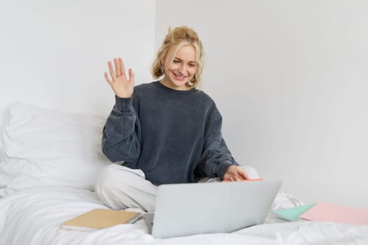Portrait of young happy woman, connects to video chat, using laptop, waving hand at camera, saying hello to someone online, sitting on bed, studying, e-learning or working from home.