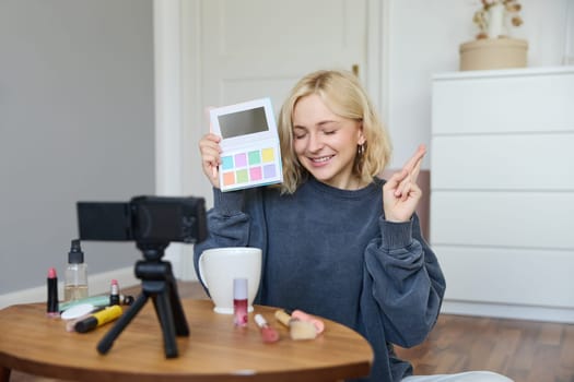 Portrait of beautiful lifestyle blogger, girl records a video on her camera for social media, shows palette of eyeshadows, does a makeup tutorial for her followers, sits in her room.